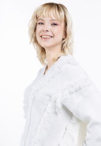Biały sweter z miękkim, teksturowanym wykończeniem wzdłuż brzegów, z dekoltem w kształcie litery V i lekkim materiałem. Modelka uśmiechająca się w profilu.