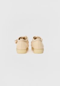 Scarpe in tela beige con tacco basso, punta arrotondata e piccolo fiocco sul retro. Il tessuto testurizzato e la suola in gomma offrono aderenza.