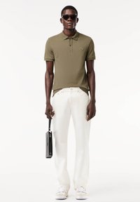 Olíva zöld polo póló, slim fit, rövid ujjak, hímzett logó. Fehér nadrággal párosítva, fekete táskát tart, sötét napszemüveget visel.