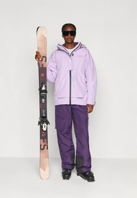 Person trägt eine lila Skijacke, dunkellila Hosen, schwarze Skischuhe und große schwarze Sonnenbrille und hält ein aufrechtes Ski mit Bindungen.