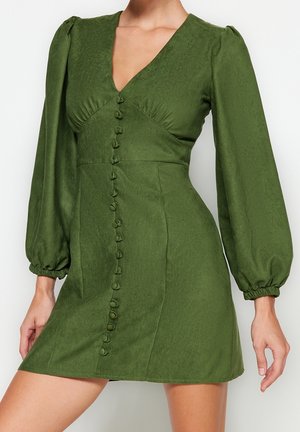 Frau trägt ein eng anliegendes olivgrünes Kleid mit gepufften langen Ärmeln, einem tiefen V-Ausschnitt und einer Reihe mit Stoff bezogener Knöpfe vorne.