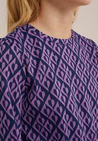 Lila och marinblå mönstrad tyg med hjärtformer inom ett geometriskt mönster, mjuk textur och rund halsringning.