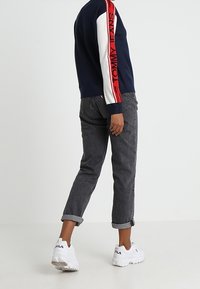 Pull bleu marine avec des accents rayés blancs et rouges, jean gris retroussé et baskets blanches avec logo noir. Tenue décontractée vue de dos.