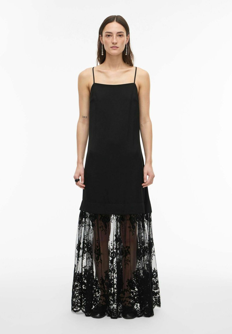 Femme portant une robe noire sans manches avec une superposition de jupe en dentelle florale transparente, debout devant un fond blanc uni.