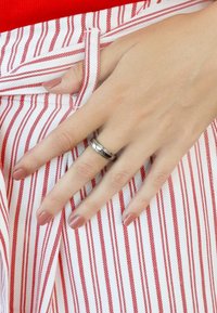 Zilveren ringen met een gedraaide ontwerp en lineaire accenten, gedragen aan een hand die rust op een rood-wit gestreepte stof. De nagels zijn neutraal van kleur.
