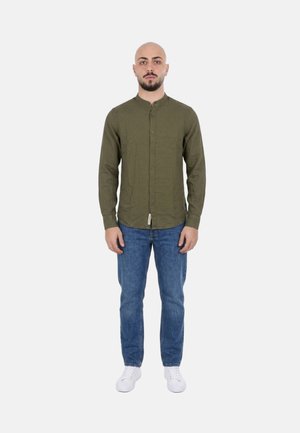 Uomo in piedi rivolto verso di noi che indossa una camicia verde oliva con bottoni, jeans blu e scarpe da ginnastica bianche su uno sfondo chiaro uniforme.