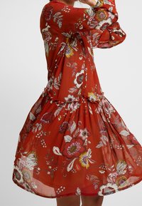 Femme portant une robe florale rouge fluide avec des fleurs roses, blanches et jaunes, tournant pour montrer le mouvement et les détails du tissu de la robe.