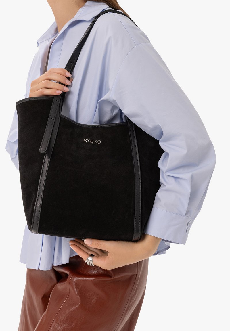Femme portant une chemise bleu clair et un pantalon marron tient un sac à main en daim noir Rylko avec des sangles en cuir à l'épaule.