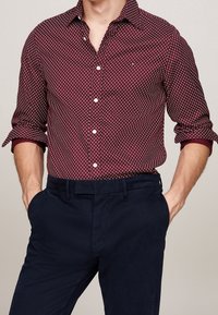 Bordeaux-gemusterde shirt met witte diamanten, lange mouwen opgerold, gecombineerd met donkerblauwe broek, gestructureerde stof en knoopdetails.