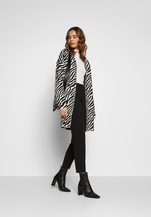 Cardigan long à motif zèbre en noir et blanc, associé à un pantalon noir ajusté et des bottines noires à talons. Tissu lisse et texturé.
