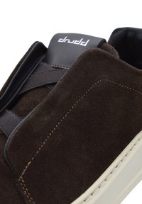 Brown suede sneaker with elastic strap, white rubber sole, and black leather accents. Notable stitching detail along the side.