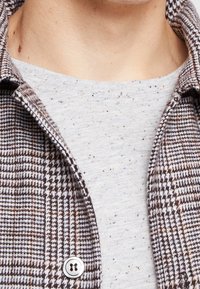 Zblízka je zachycena osoba v hnědo-bílém kostkovaném saku přes světle šedou tečkovanou košili.