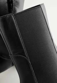 Schwarze Stiefeletten mit einem Obermaterial aus glattem Leder und einem strukturierten, dehnbaren Stoffeinsatz. Seitlicher Reißverschluss für ein einfaches An- und Ausziehen.