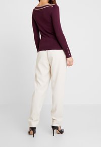 Burgundin pitkähihaisen neuleen, jossa on hieman kaareva pääntie ja beige yksityiskohta. Hihassa on hopeiset napit. Yhdistetty kermanvärisiin housuihin.