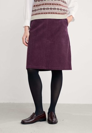 Frau, die einen lila Cord-Knielangen Rock, eine gemusterte Strickweste, schwarze Strumpfhosen und dunkelbraune Loafers trägt, steht mit einer Hand in der Tasche.