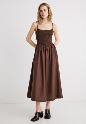 Junge Frau mit blonden Haaren, die ein braunes, ärmelloses Midikleid mit gerafftem Oberteil und schwarze Mules mit Blockabsatz trägt, steht vor einer weißen Wand.