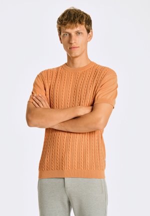 Pomarańczowy sweter z krótkim rękawem, pleciony, z wzorem wypukłych pionowych kabli, okrągłym dekoltem i ściągaczem. W połączeniu z szarymi spodniami.