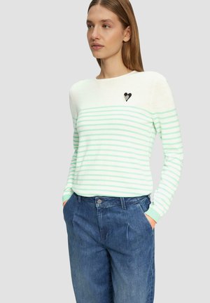 Pull rayé vert clair et blanc à manches longues, col rond, avec un patch en forme de cœur noir sur la poitrine, associé à un pantalon en denim bleu.