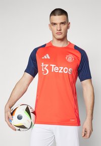 Sarkans un zils īsu piedurkņu futbola kreklis ar apaļu kaklu, kas satur sponsoru logotipus un Mančestras United emblēmu. Viegs audums, plāns piegriezums.