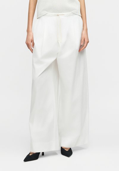 Pantalon blanc à jambes larges en tissu texturé, avec des plis et une ceinture élastique, associé à des talons noirs à bout pointu.