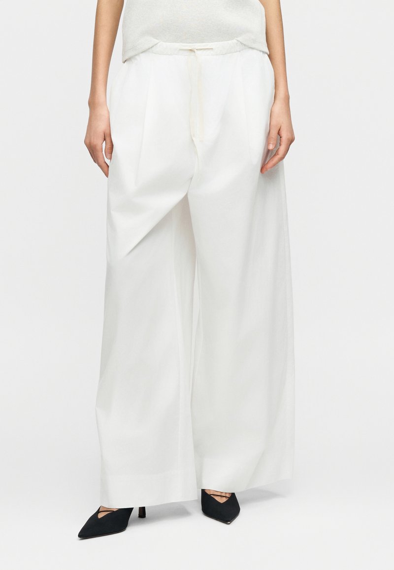 Λευκό παντελόνι με φαρδύ παντελόνι από υφή υφάσμ ατος, με πιέτες και ελαστική μέση, συνδυασμένο με μαύρα τακούνια με μυτερή μύτη.