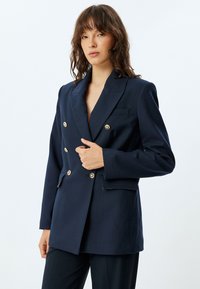 Sininen tupla-painikkeinen bleiseri, joka on valmistettu teksturoidusta kankaasta. Siinä on kultasävytteiset napit, viistosakset ja yksi rintatasku, jossa on pinstripe-kuvio.