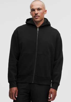 Mann mit rasiertem Kopf, der einen schwarzen Kapuzenpullover mit Reißverschluss und Vorderstaschen trägt, steht vor einem schlichten hellgrauen Hintergrund.