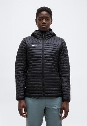 Άτομο που φοράει μαύρο μπουφάν Mammut με καπιτονέ σχέδιο και κουκούλα, στέκεται μπροστά σε απλό ανοιχτό γκρι φόντο, με τα χέρια χαλαρά στο πλάι.