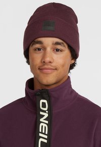 Maroon beanie with ribbed texture and black logo patch; purple fleece jacket with a zip and contrasting black zipper detailing.