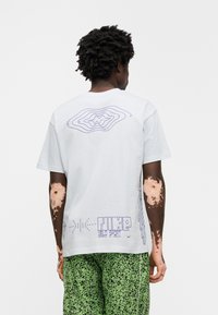 Λευκό κοντομάνικο μπλουζάκι με μοβ γραφιστικό σχέδιο στην πλάτη, που περιλαμβάνει το κείμενο "NIKE VELO SPORT" και αφηρημένα σχήματα.