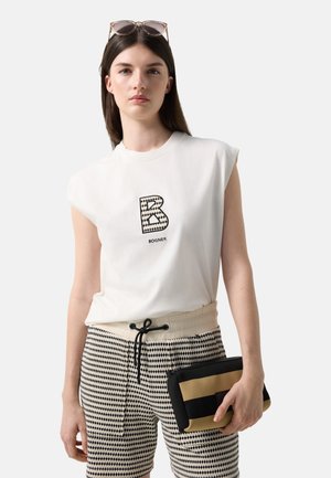 Jonge vrouw met lang donker haar, dragen een wit mouwloos topje met een patroon van het letterlogo "B", gemêleerde korte broek, houd een zwarte en beige clutch vast.