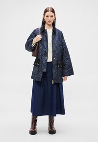 Gewatteerde marineblauwe jas met gouden accessoires, zakken aan de voorkant en een zachte kraag, gecombineerd met een crèmekleurige trui, denim rok en bruine laarzen.