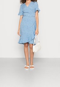 Robe enveloppante bleue avec imprimé floral blanc, manches volantes et ourlet. Associée à des sandales blanches à enfiler et un petit sac à main blanc.