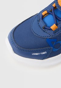 Blauer Sportschuh mit einer strukturierten Stoffoberfläche, orangefarbenen Akzenten und einer weißen Sohle. Mit "LOONEY TUNES"-Logo an der Seite.