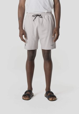 Persoon draagt lichtgrijze, getextureerde shorts met een zwarte trekkoord, wit t-shirt en zwarte sandalen met twee banden, staand op een witte achtergrond.