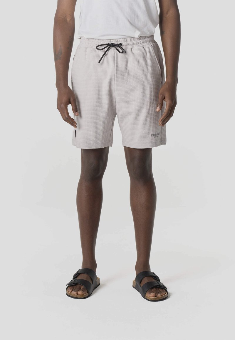 Persoon draagt lichtgrijze, getextureerde shorts met een zwarte trekkoord, wit t-shirt en zwarte sandalen met twee banden, staand op een witte achtergrond.