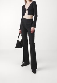 Haut noir cropped à manches longues avec un détail anneau à l'avant, associé à un pantalon évasé noir taille haute. Le modèle tient un sac à main texturé noir.