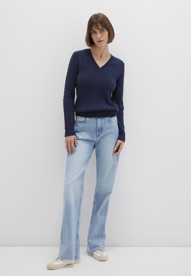 Marineblauer Kabelstrickpullover mit V-Ausschnitt, kombiniert mit hellblauen Schlagjeans und beigen Schuhen, vor einem schlichten weißen Hintergrund platziert.