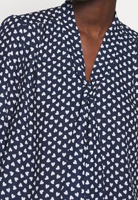 Blusa blu navy caratterizzata da un motivo a cuori bianchi, con scollatura a V e dettaglio con fiocco sul davanti. Tessuto leggero e morbido.
