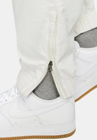 Vita atletiska byxor med siddragkedjor, matchade med vita sneakers och grå strumpor. Byxorna har elastiska muddar och en mjuk yta.