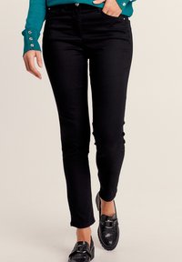 Jean noir ajusté avec une texture lisse, doté de cinq poches et de quincaillerie en métal, associé à des mocassins noirs et un haut teal avec des accents de boutons.