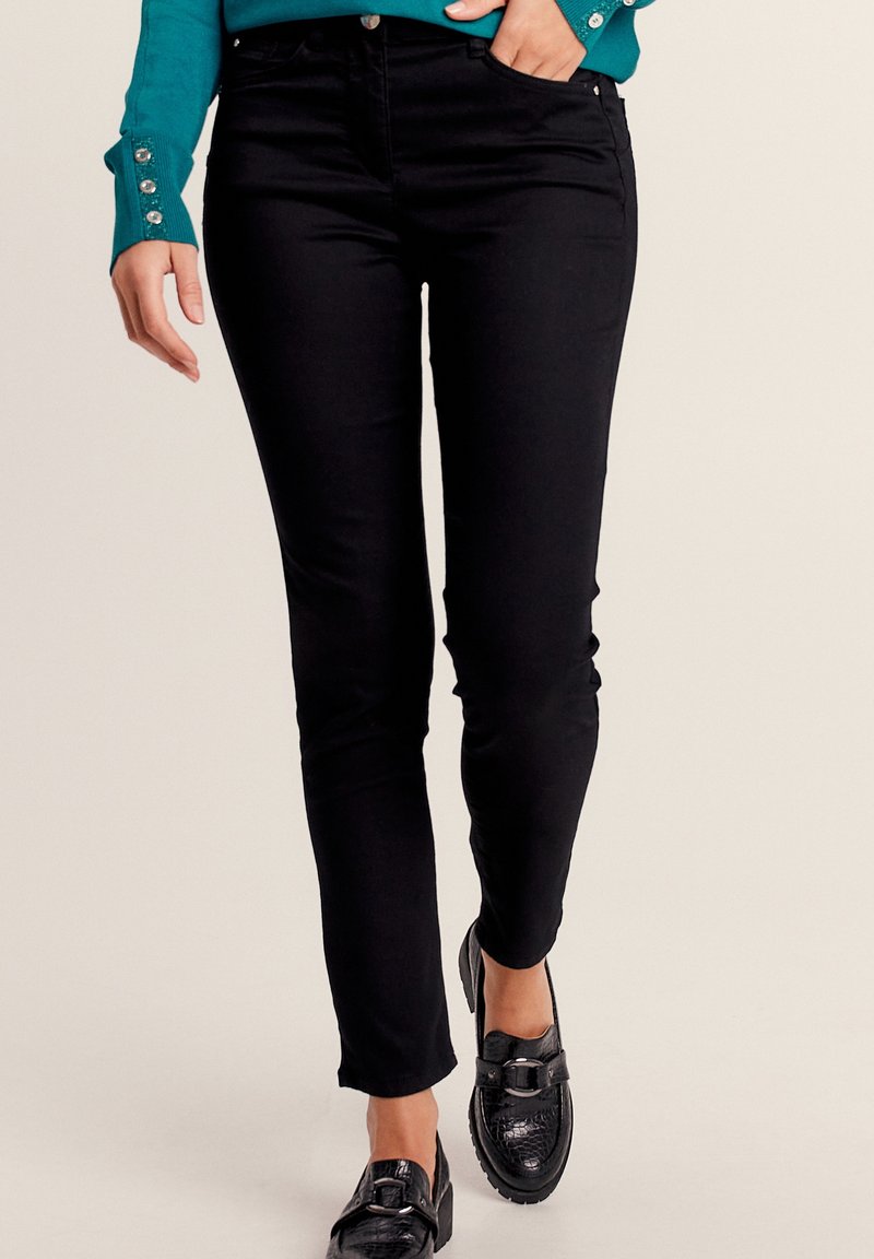 Jean noir ajusté avec une texture lisse, doté de cinq poches et de quincaillerie en métal, associé à des mocassins noirs et un haut teal avec des accents de boutons.