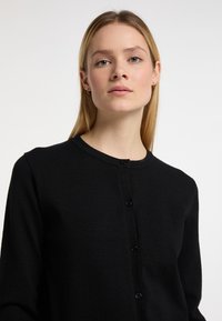 Cardigan negru fabricat din material moale, având un decolteu rotund, mâneci lungi și un închidere cu buton pe față.
