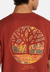 Camiseta de algodón color óxido con un gráfico circular de un árbol con ramas intrincadas y escenas de la naturaleza en amarillo, verde y naranja.