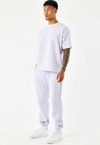 Camiseta corta de manga corta en color púrpura claro con un pequeño logotipo blanco, combinada con pantalones jogger a juego que tienen tobillos ajustados y un detalle de logotipo. Zapatillas blancas.