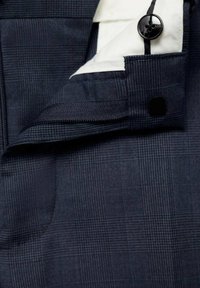 Gros plan d'un tissu de costume à carreaux bleu foncé montrant la ceinture avec un bouton noir et une doublure intérieure blanche.