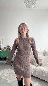 Jeune femme aux cheveux blonds portant une robe marron en maille torsadée et des bottes noires, debout dans un salon confortable avec des meubles blancs.