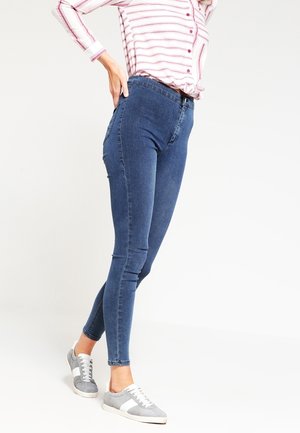 Vrouw die een hoge taille blauw skinny jeans draagt, grijs-witte sneakers en een wit shirt met rode strepen, staand met één hand op haar heup.