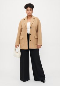 Beige oversize blejzr s dvěma předními kapsami, černými knoflíky a hladkým povrchem, doplněný širokými černými kalhotami a bílou kabelkou.