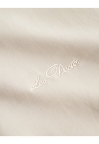 Lys beige stof med en glat tekstur, der har en delikat hvid broderi af "Lis Dea." Simpelt, elegant design med en subtil glans.